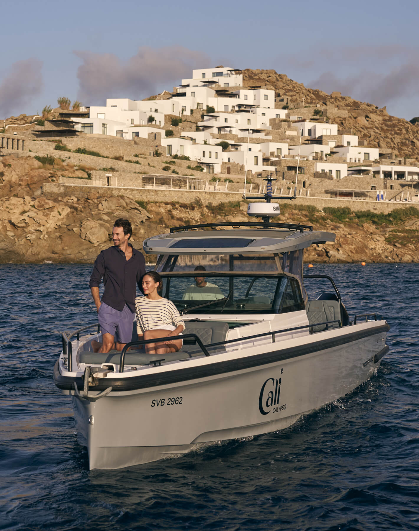 A serene boat ride near Cali, featuring a couple aboard one of Cali's sleek vessels as they pass by the stunning hillside resort of Cali on Mykonos. The early evening light bathes the scene in a golden hue, showcasing the idyllic coastal landscape.