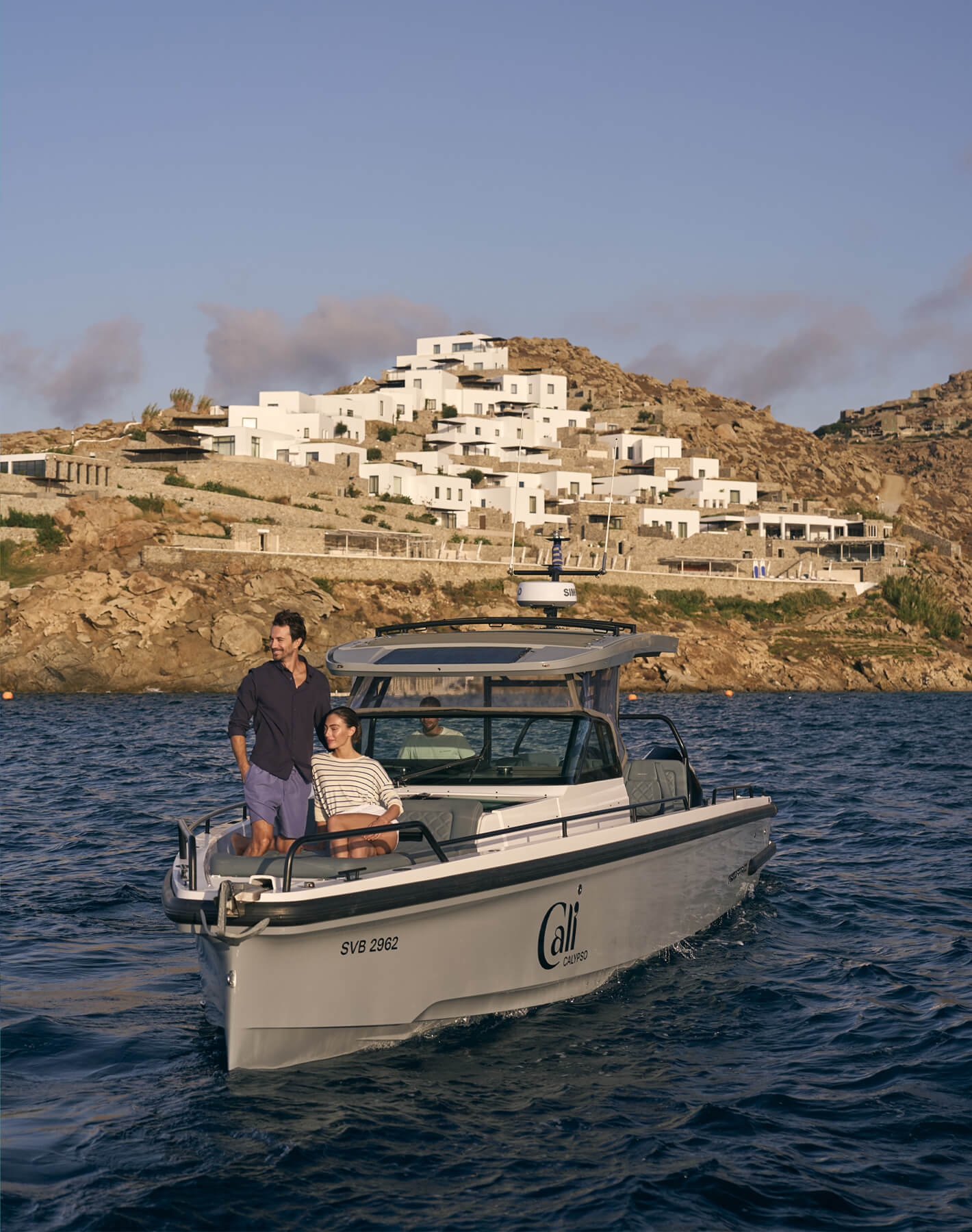 Luxury resort experience at Cali Mykonos: couple on Axopar boat ride across the Aegean Sea with Cali in the background, Mykonos Greece.