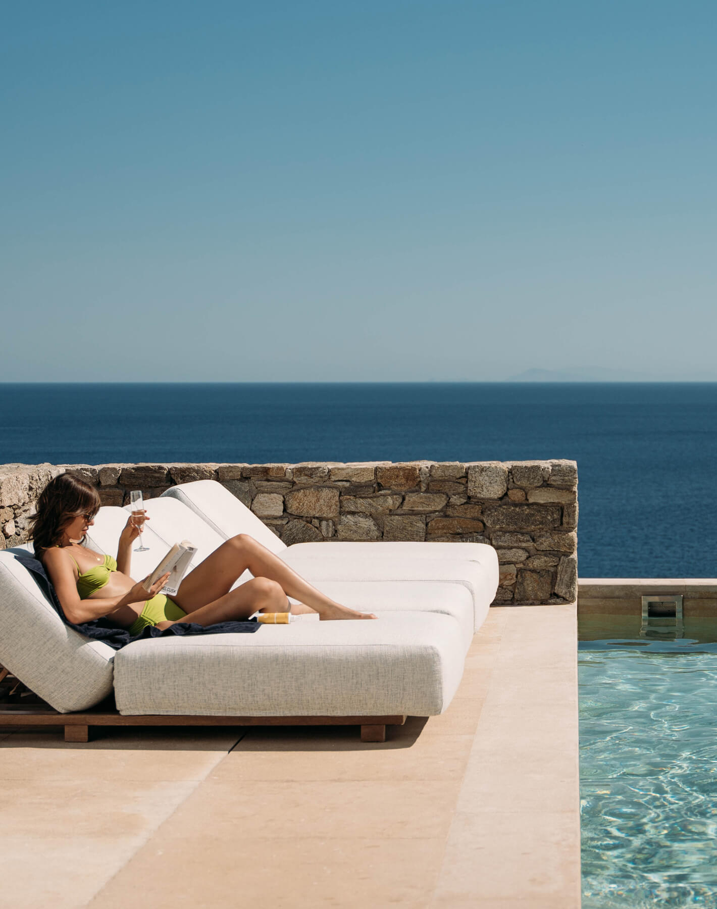 Peaceful afternoon at Cali Mykonos — woman sunbathing on a terrace by her villa’s private pool, reading a book with breathtaking Aegean Sea views.