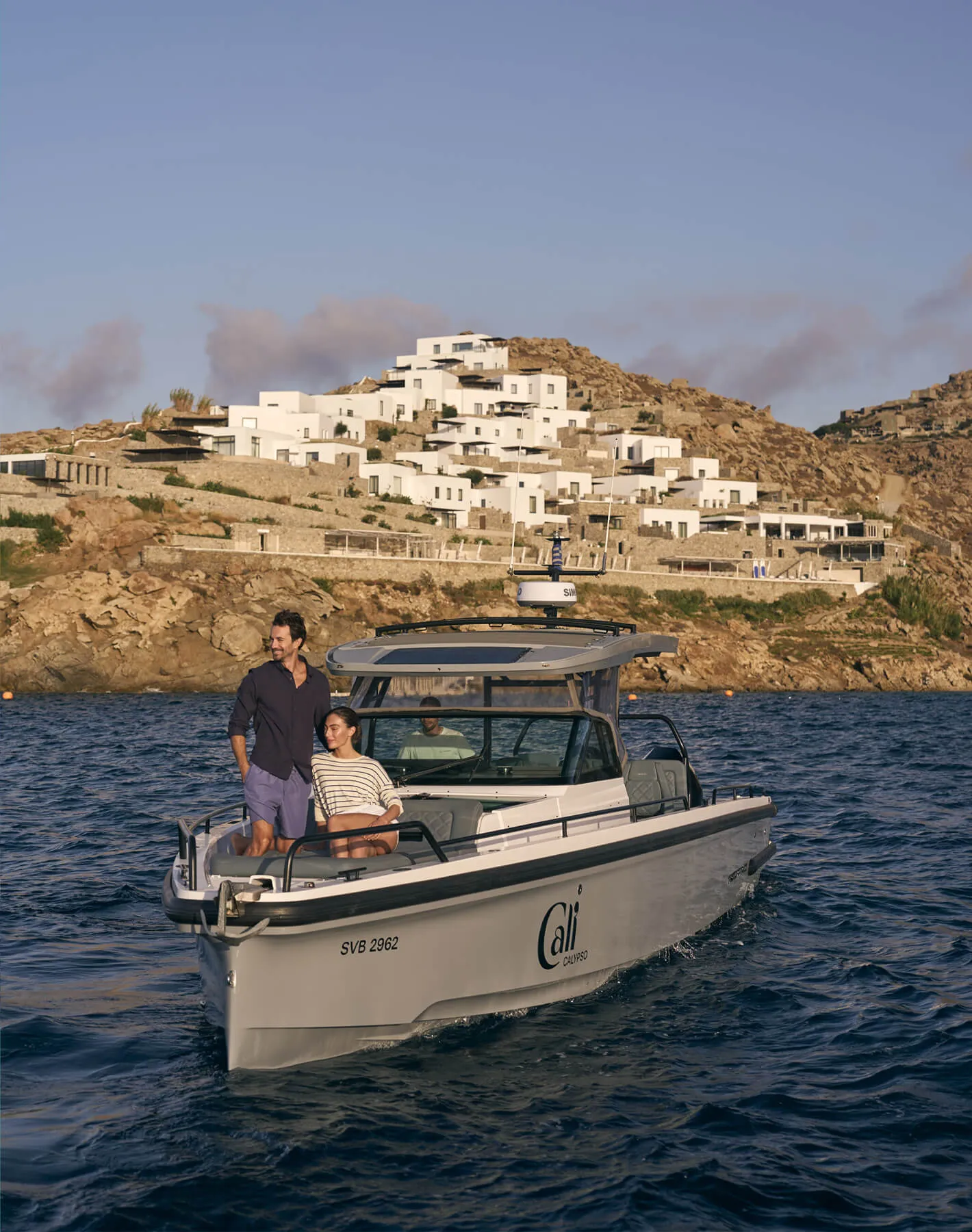 Luxury resort experience at Cali Mykonos: couple on Axopar boat ride across the Aegean Sea with Cali in the background, Mykonos Greece.