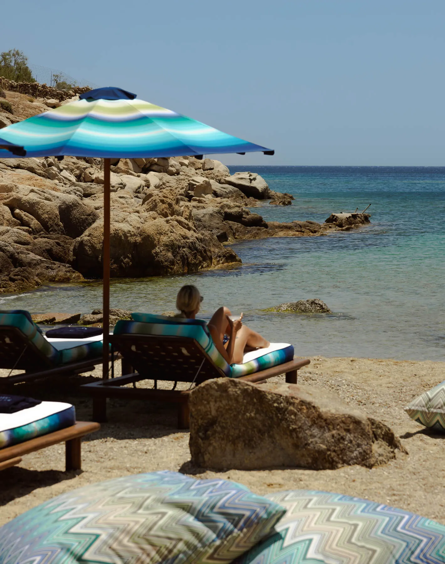 Luxury beach experience at Cali Mykonos resort in Greece — guest relaxing by the Aegean Sea with sunlight reflecting on the water.