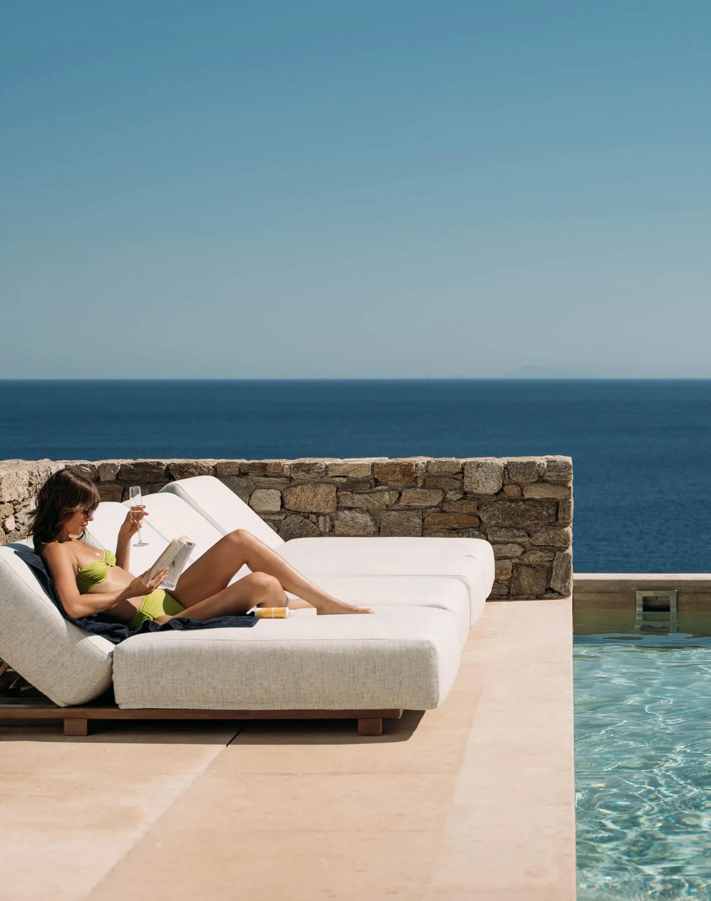 Peaceful afternoon at Cali Mykonos — woman sunbathing on a terrace by her villa’s private pool, reading a book with breathtaking Aegean Sea views.